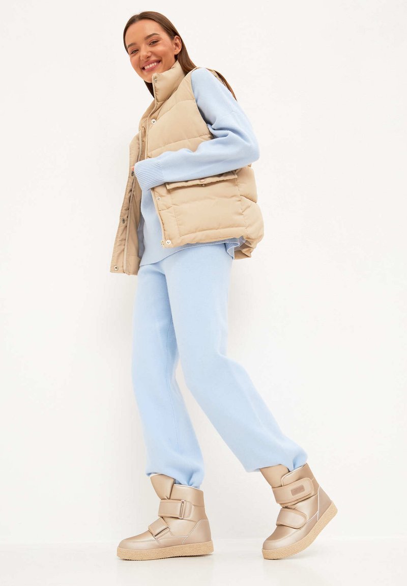 Bézs színű puffertakaró mellény világoskék pulóver és hozzá illő melegítőnadrág felett. Masszív bézs színű, magas szárú sportcipők két pánttal. Simá anyagok és laza fazon.