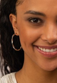 Goldfarbene metallene Creole mit einem gedrehten Design, das eine glatte, reflektierende Oberfläche zeigt. Sitzt eng am Ohr.