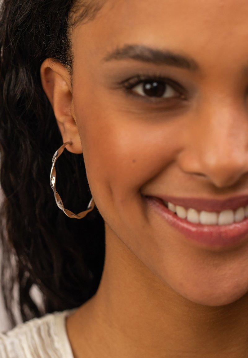 Goldfarbene metallene Creole mit einem gedrehten Design, das eine glatte, reflektierende Oberfläche zeigt. Sitzt eng am Ohr.