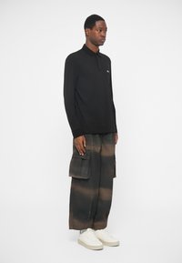 Camisa polo preta de manga longa com colarinho; combinada com calças cargo oversized em tons escuros e salpicos claros; ténis brancos.