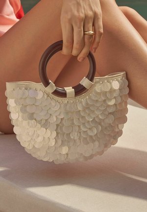 Mână care ține o poșetă bej acoperită cu paiete, cu mânere rotunde din lemn, odihnindu-se pe picioare goale încrucișate, pe un fundal neutru.