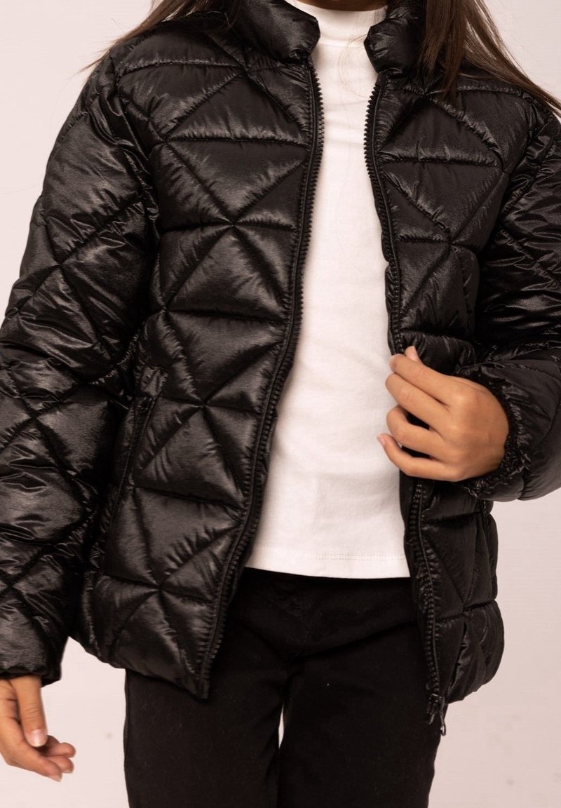 Kind, das eine schwarze gesteppte Jacke über einem weißen Shirt und schwarzen Hosen trägt, während es mit einer Hand den Reißverschluss anpasst.