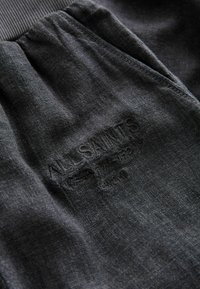 Tissu gris foncé avec taille élastique froncée et texte embossé indiquant "ALLSAINTS" et des mots plus petits en dessous.