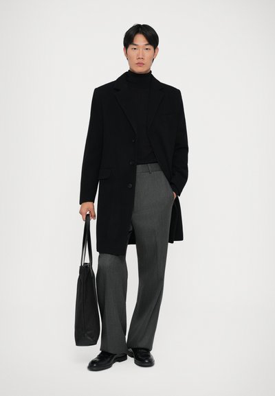 Cappotto lungo di lana nera, dolcevita nero, pantaloni grigi a gamba larga. Il modello tiene una borsa nera; scarpe eleganti nere lucide completano l'outfit.