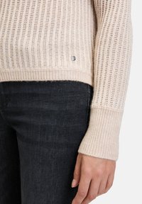 Beige gerippter Pullover mit strukturiertem Strick, der ein silbernes Logodetail am Saum und kontrastierende gerippte Bündchen aufweist, kombiniert mit dunklen Jeans.