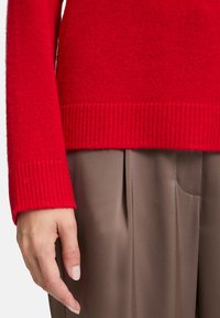 Dłoń z neutralnym lakierem do paznokci obok rękawa czerwonego swetra i brązowych spodni plisowanych na jasnym tle.