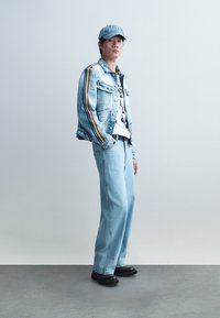 Joven con chaqueta de mezclilla azul claro con rayas de arcoíris en las mangas, jeans a juego, camiseta gráfica, gorra azul y zapatos negros, de pie.