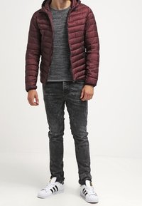 Doudoune bordeaux avec un motif matelassé horizontal, pull en tricot gris, jean déchiré gris foncé et baskets blanches Adidas.