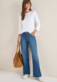 Hvid langærmet top, blå brede jeans og hvide sneakers. Holder en brun ruskinds skuldertaske. Enkelt design og afslappet pasform.