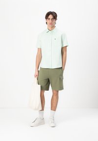 Lys grønne kortermede skjorte med knapper, olivengrønne cargo shorts, hvide sneakers og en krämfarvet tote bag. Har logo på skjorte og shorts.