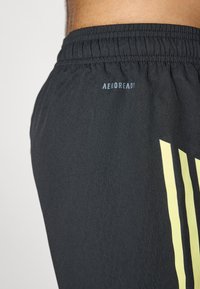 Schwarze Sportshorts mit elastischem Bund. Mit einem hellgelben Streifenmuster und dem Text "AEROREADY" in der Nähe des Bundes.