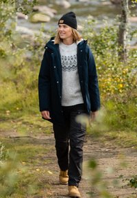 Niebieska ocieplana kurtka, jasnoszary sweter w wzory, czarne spodnie, beżowe buty. Naturalne otoczenie z zielonymi liśćmi i rzeką w tle.