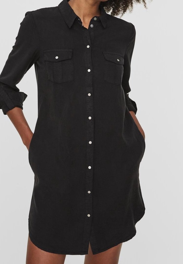 Femme portant une robe-chemise noire à manches longues boutonnée, manches retroussées et deux poches poitrine, mains dans les poches latérales.