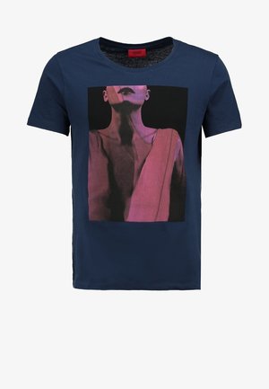 Homme portant une veste en cuir marron foncé, un t-shirt graphique bleu marine, un jean noir et des bottes noires, se tenant les jambes croisées.