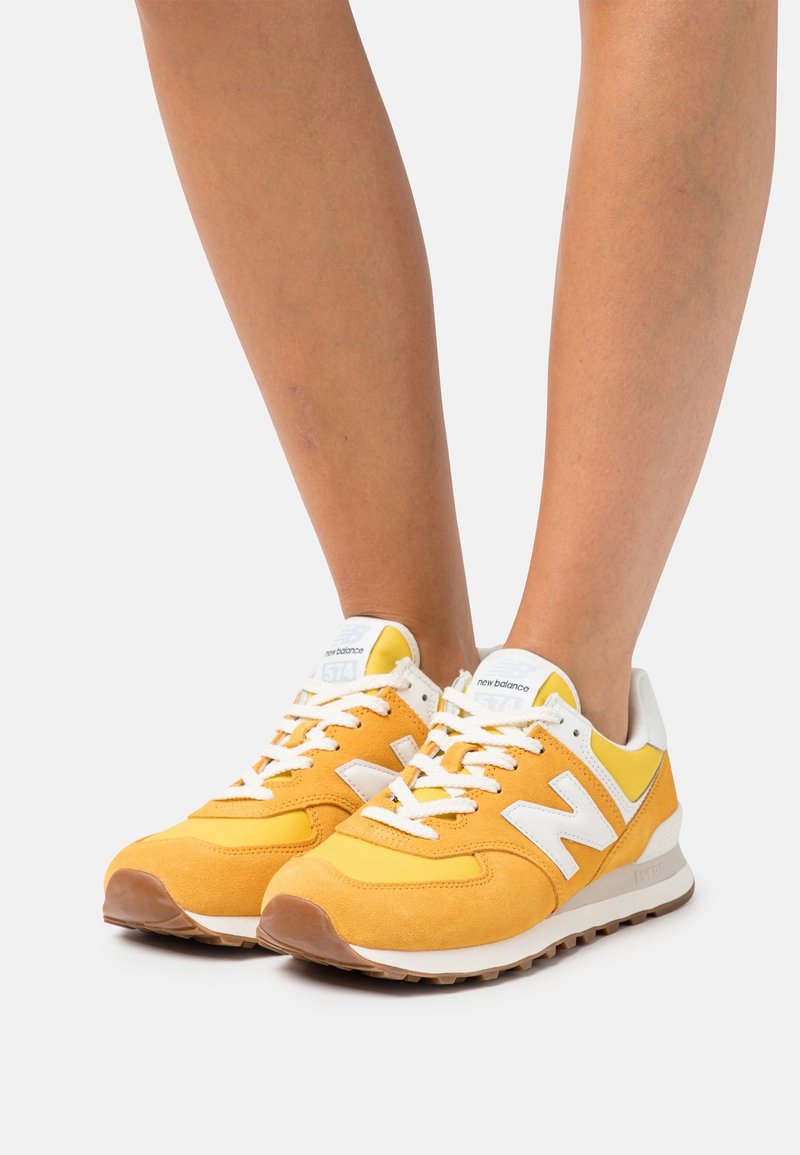 Personne portant des baskets New Balance jaunes et blanches avec des lacets blancs, debout sur une surface unie gris clair.
