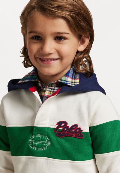 Sudadera a rayas blancas y verdes con capucha azul marino, que presenta un logo bordado y un diseño geométrico. Debajo, se puede ver una camisa de cuadros.
