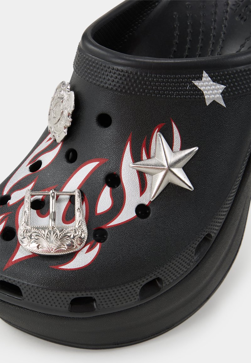Zuecos negros con patrón de llamas; adornados con hebilla plateada, estrella y emblema. Fabricados en material texturizado; cuentan con agujeros de ventilación.
