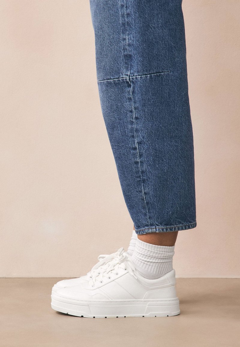 Baltos sportbačiai su tekstūruoto audinio viršumi ir gumine padais, derinami su baltais, su ažūriniais raštais kojinaitėmis, rodomi kartu su trumpintais mėlynais džinsais.