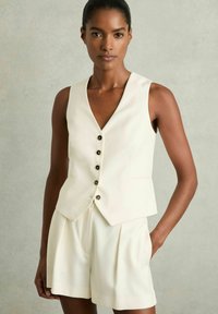 Gilet aderente senza maniche color crema con tre bottoni, abbinato a pantaloni corti plissettati coordinati. Texture liscia e design minimalista.