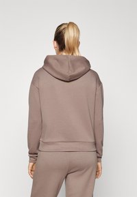 Sudadera con capucha en tela marrón con un ajuste holgado. Cuenta con puños y dobladillo acanalados, textura suave y un bolsillo tipo canguro.