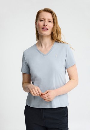 Vrouw met lang rood haar, die een lichtblauw T-shirt met V-hals en korte mouwen en zwarte broek draagt, staand tegen een eenvoudige witte achtergrond.