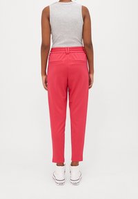 Personne portant un pantalon taille haute rose vif, un haut gris sans manches et des baskets montantes blanches Converse, debout dos à l’appareil sur un fond uni.