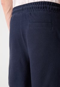 Pantalones de chándal azul marino con un tejido texturizado, cintura elástica y un detalle de bolsillo trasero, mostrados desde un ángulo lateral.