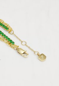 Bracciale dorato con gemme verdi, dotato di chiusura e di un'etichetta incisa. Il design include catene collegate e una finitura texture.