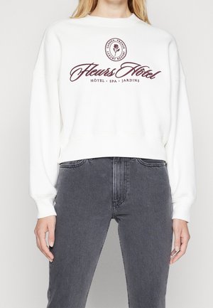 Vit sweatshirt med rund halsringning, långa ärmar och en logo i vinröd text. Kombinerad med högmidjade grå jeans. Slät textur.