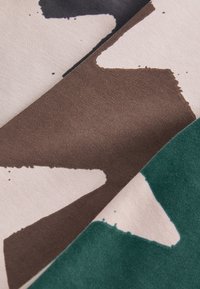 Gros plan d’un tissu plié avec des formes abstraites en marron foncé, vert, noir et blanc sur un fond beige.