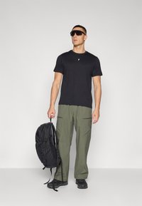 Black t-shirt, olive green cargo pants, and a black backpack. Accessories include dark sunglasses. Casual outdoor apparel.