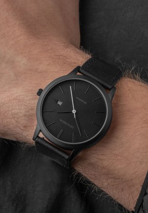 Black analog wristwatch with a matte black circular case, black leather strap, minimalist dial, and silver hour/minute markers.