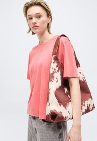 Jauna sieviete, tērpusies koraļļrozā T-kreklā un pelēkās džinsās, pār plecu nēsā lielu brūnu un baltu plecu somu ar rakstu.