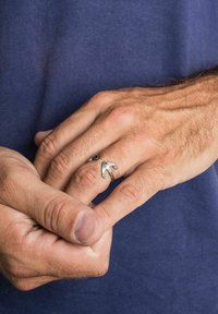 Anello in argento con un design che ricorda la testa di un uccello, posizionato sopra la mano di una persona su uno sfondo di tessuto blu navy. Superficie texturizzata visibile.
