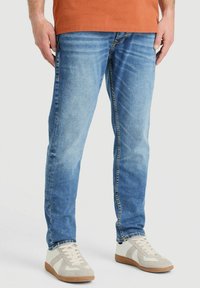 Pantalones vaqueros de mezclilla azul claro con ajuste slim, textura desgastada, diseño tradicional de cinco bolsillos, combinados con zapatillas blancas y beige.