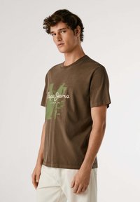 Camiseta de algodón marrón con un diseño gráfico verde que incluye el texto "Pepe Jeans London"; escote redondo, mangas cortas y un corte relajado.