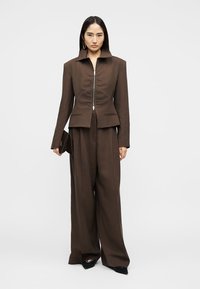 Femme portant un blazer zippé marron sur mesure avec un pantalon large assorti, tenant une pochette marron, debout devant un fond uni.
