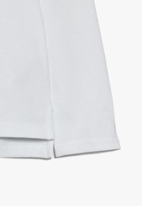 Bílá texturovaná látka s bočním rozparkem a dvakrát prošitým lemem na oděvu, zobrazená na bílém pozadí.