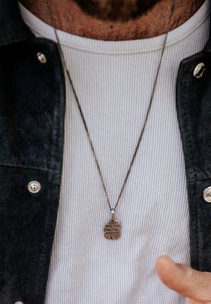 Silver pendant necklace with engraved text on a chain, worn over a ribbed white shirt and a dark jacket. Text reads, "REAL EYES REALIZE REAL LIES."