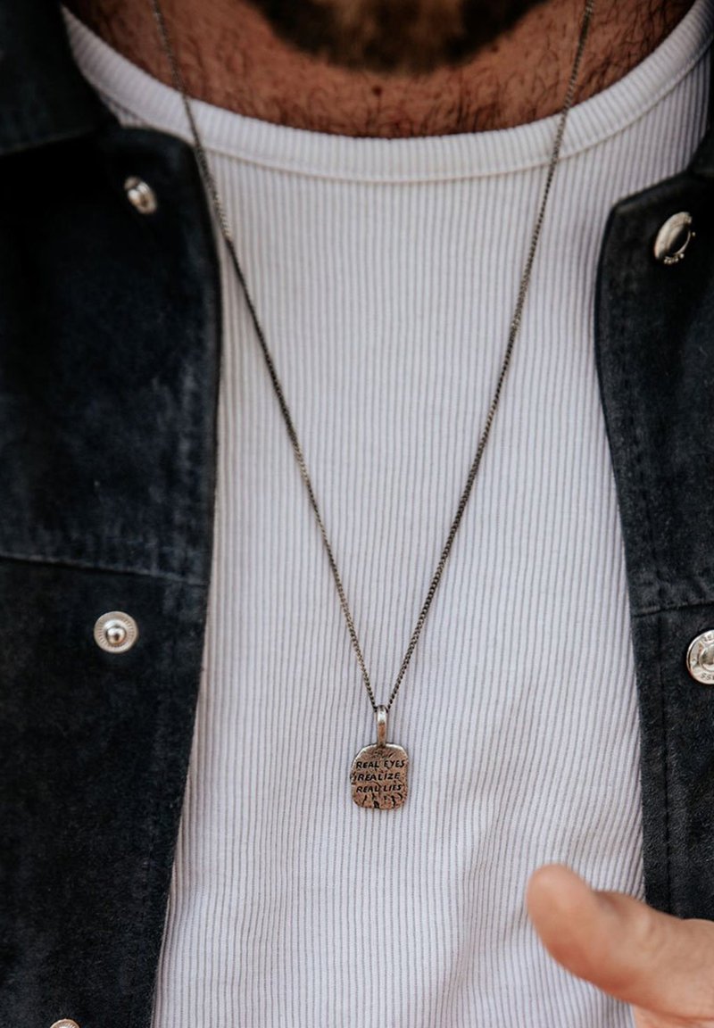 Collana con pendente in argento con testo inciso su una catena, indossata sopra una maglietta bianca a coste e una giacca scura. Il testo recita: "GLI OCCHI REALI RICONOSCONO INGANNO REALE."