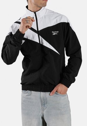 Mann trägt eine schwarz-weiße Reebok Zip-up-Jacke und hellblaue Jeans, hält den Reißverschluss nahe am Hals mit einer Hand.