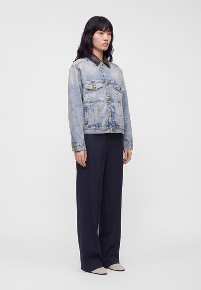 Femme aux cheveux noirs portant une veste en jean clair avec des boutons dorés, un pantalon large noir et des chaussures pointues beiges sur fond blanc.