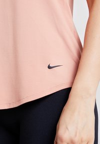 T-shirt de sport Nike rose clair avec une texture lisse, présentant un petit logo noir à l'ourlet. Associé à des leggings noirs.