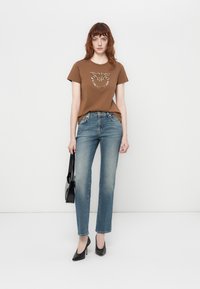 T-shirt marron avec un design en perles et sequins, associé à un jean délavé moyen. Le mannequin tient un sac à main noir et porte des chaussures à talons noirs.