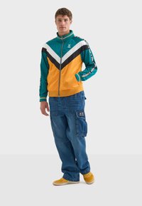 Färgglad tracksuitjacka i teal, orange och svart med ett chevronmönster; kombinerad med blå cargobyxor och gula sportskor.