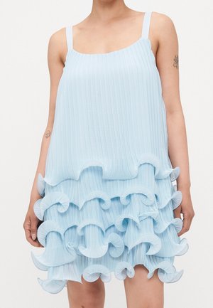 Robe sans manches bleu pâle plissée avec plusieurs volants superposés sur la jupe, portée par une personne avec des tatouages visibles sur les bras.