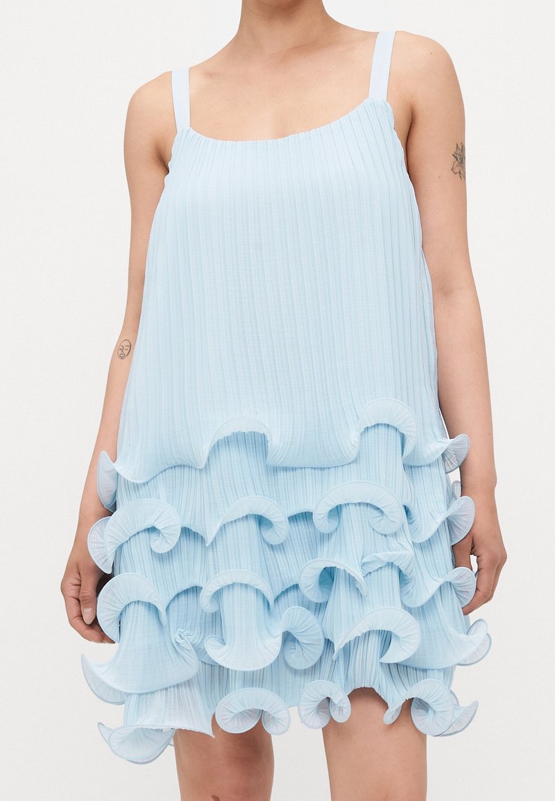 Robe sans manches bleu pâle plissée avec plusieurs volants superposés sur la jupe, portée par une personne avec des tatouages visibles sur les bras.