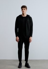 Czarna bluza z zamkiem na suwak na czarnej koszulce, zestawiona z czarnymi, zwężanymi spodniami i czarnymi trampkami. Gładki materiał, minimalistyczny design.