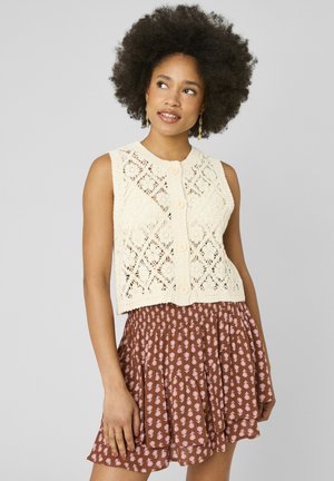 Femme aux cheveux afro naturels portant un gilet tricoté crème sans manches boutonné et une jupe marron à motifs à étages, regardant légèrement vers le haut et à droite.