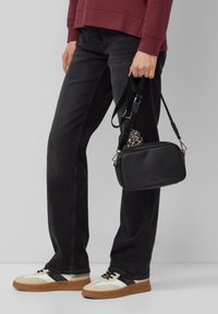 Borsa nera con texture, dotata di accessori in argento, abbinata a jeans scuri e sneakers con accenti beige e una striscia nera.
